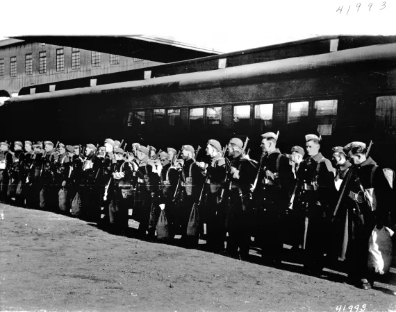 Image is a black-and-white photograph showing the first Canadian contingent of troops standing at attention before a train car.