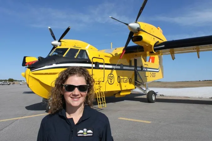 A young woman wearing sunglasses smiles as she looks towards the camera. A large yellow aircraft is visible on the runway behind her; the word “Ontario” is clearly visible on the side of the aircraft.