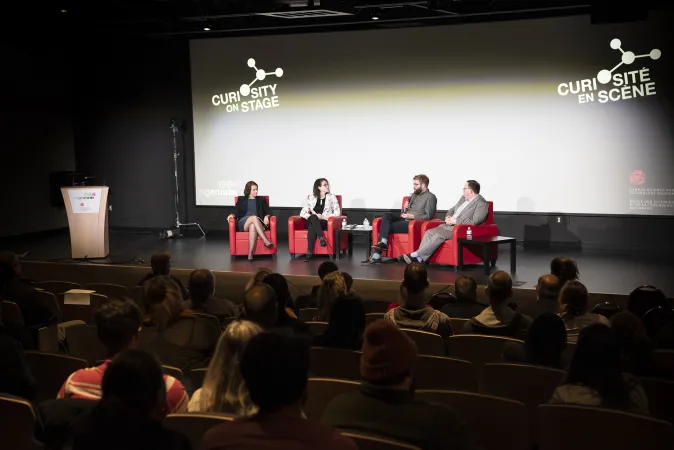 Four people sitting on a stage at the front of a large room. They are sitting on individual bright red futons and one of them is talking into a mic. Behind them, a powerpoint presentation is projected onto a large screen that spans from floor to ceiling. The words Curiosity on Stage and Curiosite en scene with the logo is displayed at the top of the screen. There is a podium on the side of the stage.
