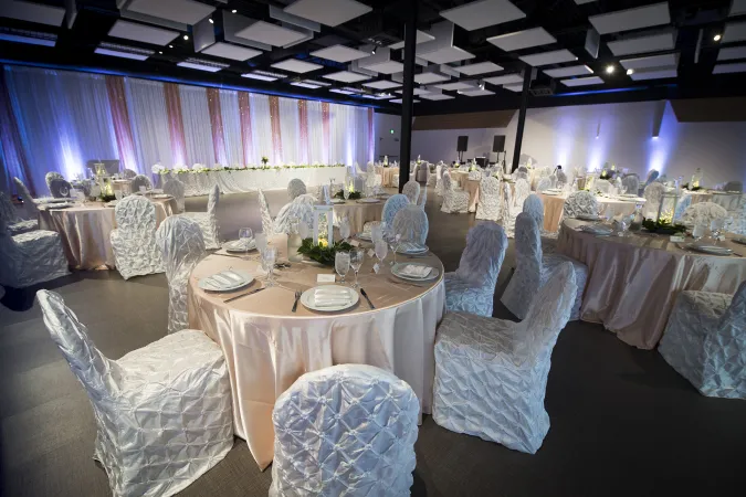 A large room with white and pink curtains and multiple round tables with floor-length pink table cloths and chairs with lush white cloth draping. The tables are set up formally for a dinner and the room is illuminated in white and blue lights.