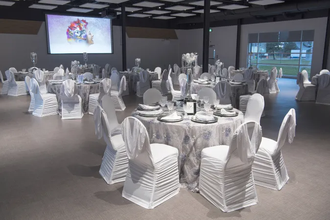 A large room with white walls and multiple round tables with floor-length white table cloths and chairs with lush white cloth draping. The tables are set up formally for a dinner and the tables have silver plates and silverware set up at each spot. There is a large projector screen on one wall of the room.