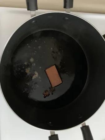 A black pot on a stove holds some black liquid that is forming small bubbles, a rectangular block of light brown sugar, and two pieces of star anise.