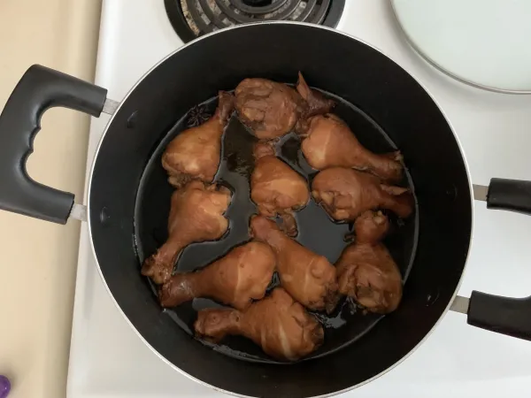 Des pilons de poulet de couleur brune sont disposés dans une casserole noire sur une cuisinière blanche. La casserole contient une petite quantité de liquide foncé.