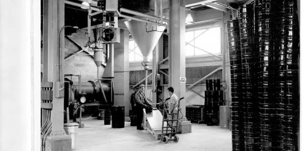 A black-and-white image shows a high-ceilinged warehouse space, with a large machine towards the back of the scene. A dolly and two men are standing next to the machine.