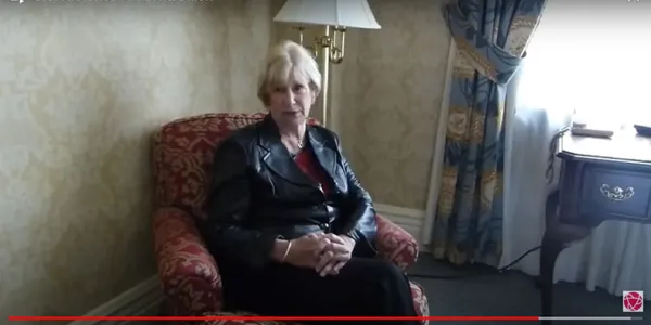 A still frame of a video featuring a woman sitting on a red armchair in the middle of a sunny room.