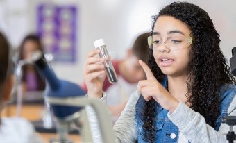 A teenage girl with long curly black hair holds a test tube half-filled with soil. The classroom behind her is out of focus.
