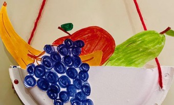 A fruit basket craft, made from a paper plate. The handcrafted basket is laced together and hung with red yarn. Handcrafted images of colourful fruits are sticking out of the top of the basket.