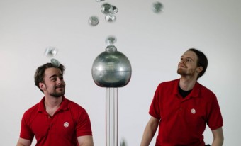 Two men in museum uniforms are on either side of a Van der Graff machine. Metallic disks float in the air, repelled by the machine.