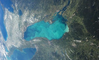 Photographie centrée sur le lac Ontario qui a une apparence turquoise trouble. L'image englobe des parties des lacs Huron et Érié le long de son côté gauche et le reste de l'image apparaît sous forme de végétation verte et de terre beige et brune.
