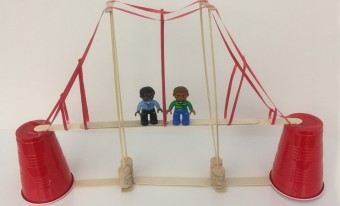 A suspension bridge made of Popsicle sticks. The Popsicle sticks sit on two upside down plastic cups on each end to raise the bridge. Wooden skewers stuck in corks serve as the posts of the bridge and ribbon acts as the suspension cables. Two LEGO people stand on the bridge.