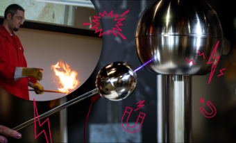 A long metal handle with a small silver ball has a spark coming out of it as it points at a Van der Graaff machine. In the top left corner there is a person in a red lab coat with protective goggles holding a stick with fire on the end of it. There are red illustrations of an explosion, a lightning bolt and a magnet on top of the image.