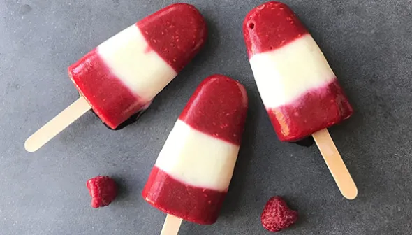 Three red-and-white striped popsicles lie on top of a grey surface, along with two strawberries. 