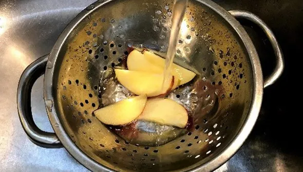 A stream of water pours over five apple slices in a metal colander.
