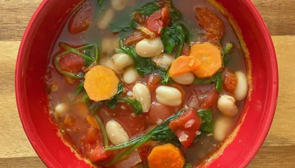 A red soup bowl sits on a wooden cutting board. The bowl is filled with soup that features spinach, tomatoes, white beans, and carrots.