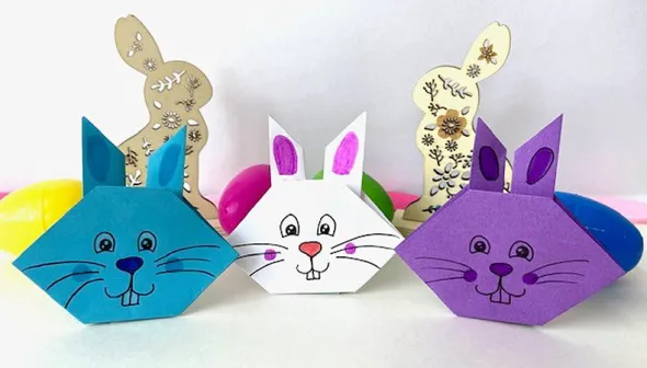 Three origami paper rabbit faces are lined up, sitting on a white background. One is blue, one is white, and one is purple; each has a cartoon-style face in black ink. Behind the paper rabbits are a collection of brightly coloured eggs and decorative wooden rabbits.