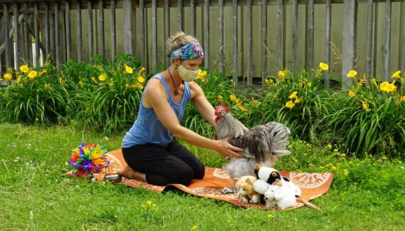 A woman wearing a face mask kneels on a yoga mat on the grass. She is reaching for a grey rooster who stands next to her on the mat; a series of toys are arranged around them. 