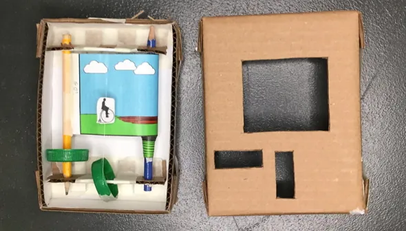 Two pieces of a cardboard craft sit on a dark surface. The left-hand piece shows the inside of a box/ pencils, strings, bottle caps, and an illustrated paper roll are visible. The right-hand piece of cardboard has cut-out holes to make a cover for the craft.