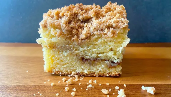 A piece of coffee cake sits on a wooden surface, against a blue backdrop.