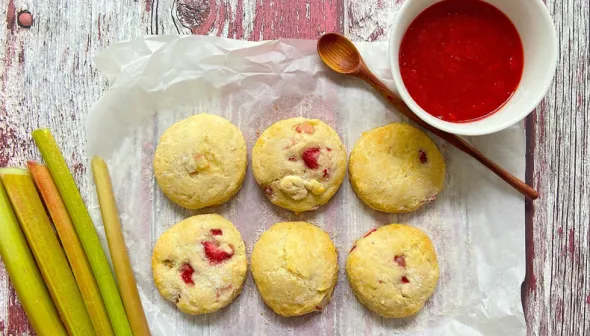 Six scones sit on a piece of wax paper against a weathered, wooden surface. Stalks of fresh rhubarb, a bowl of red jam, and a wooden spoon are visible.