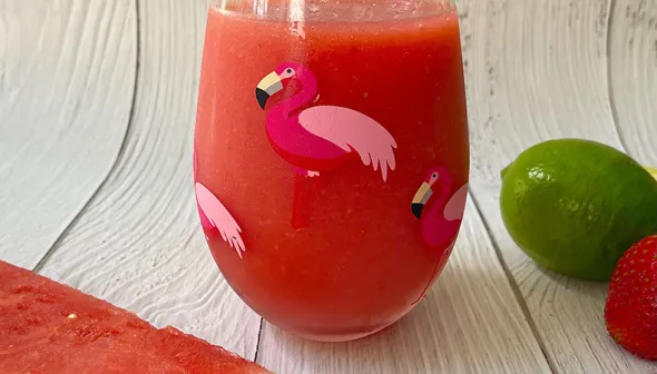 A clear glass decorated with flamingos is filled with watermelon slush. A slice of fresh watermelon and a lime sit next to the glass, against a wooden surface.