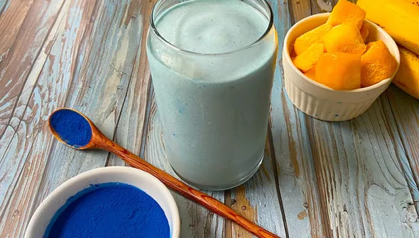 A glass filled with a thick, blue beverage sits on a rustic, wooden surface. A bowl and spoon with blue powder sits in the foreground, with mangoes and bananas in the background.  