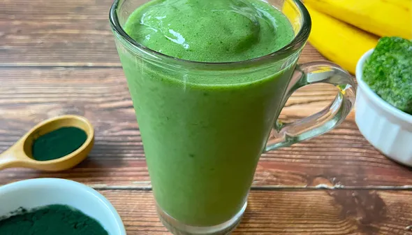 A glass filled with a thick, green beverage sits on a rustic, wooden surface. A bowl and spoon with green powder sit next to the glass, and spinach and bananas are visible in the background.
