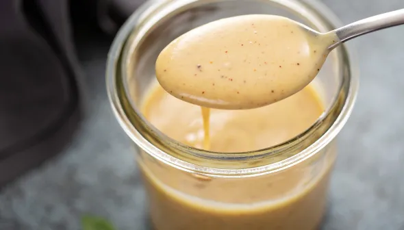 A spoon filled with honey mustard sauce is poised over the open mouth of a small jar.