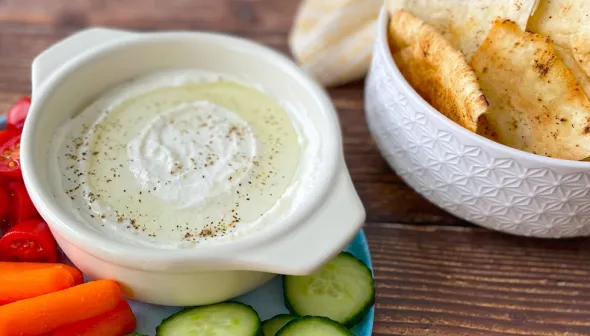Two small white bowls sit on a dark, wooden surface; one bowl is filled with feta dip and the other is filled with pita chips. Chopped carrots, peppers and cucumbers are visible around the bowl of dip.