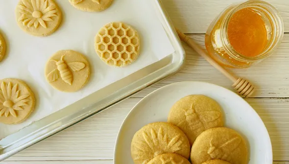 Cookies sit on a baking sheet and white plate on a white surface. Next to them, a jar of honey and honey stick are visible.  