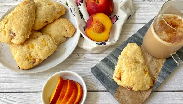An aerial view shows a plate of scones against a wooden surface. Whole and sliced peaches and a mug of coffee sit off to the side.