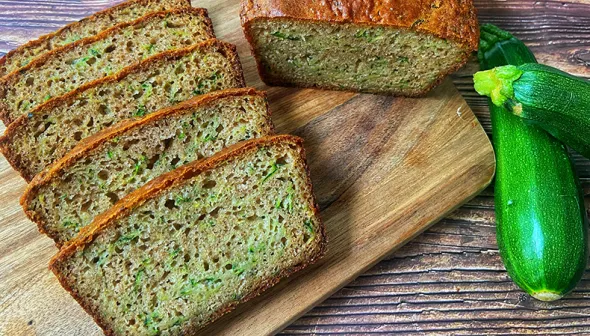 Slices of zucchini bread and half a loaf sit on a wooden platter. A zucchini lies next to the bread. 