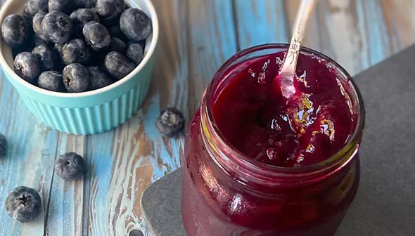 A dark purple sauce sits in an open jar, with a spoon sticking out of it. A small bowl of blueberries sits next to the jar, on top of a wooden surface.