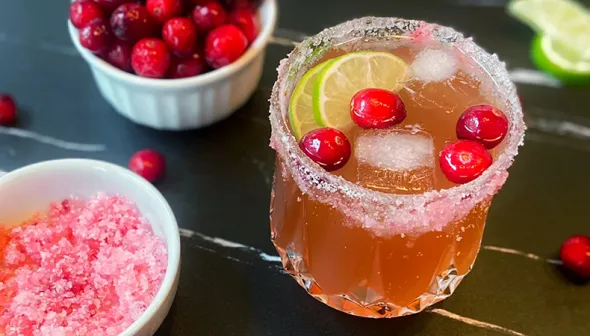 A pink-coloured drink, topped with cranberries and lime, sits on a dark wooden surface. A small bowl of cranberries and a bowl of pink sugar sit next to the glass.