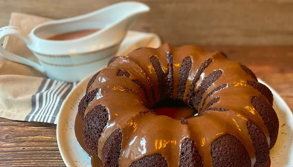 A chocolate bundt cake sits on a platter. A serving dish with chocolate sauce is visible in the background.
