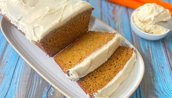 Slices of carrot cake loaf sit on a serving tray. A few carrots and frosting lie next to the loaf. 