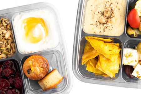 Close up of two square, clear, take-away food containers, each with four divided sections.  The container on the left has sections that feature: honey Greek yogurt, two mini-muffins, dried cranberries and pumpkin seed granola.  The container on the right is made up of: humus adorned with spices and three decorative chickpeas, seasonal vegetables, cubed feta with black and green olives and tortilla chips.