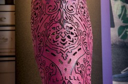 Pictured is a tightly-framed photograph of an Alleles prosthetic leg cover, as displayed in the Wearable Tech exhibition at the Canada Science and Technology Museum. The prosthetic cover's shape resembles the shape of a human leg, from knee down to ankle, but its appearance is highly stylized. It is made of a textured pink plastic, with a detailed black pattern that extends from top to bottom.