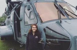 Une jeune femme sourit alors qu'elle se tient à côté d'un gros hélicoptère qui est stationné dans l'herbe.