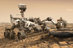 A six-wheeled rover is stationary on a light-coloured landscape. At the centre of the rover, a camera is mounted on top of a mast. A robotic arm is outstretched in front, and an instrument head at the end of the arm rests just above a rock.