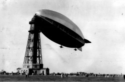 A black-and-white photograph showing a crowd gathered on the ground below the R-100, a massive blimp-like airship, tied to the top of a tall tower that looks like a lighthouse.