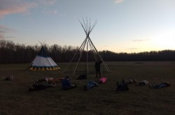 In a field, a group of people lie on the grass looking up at the sky in front of two tipis.