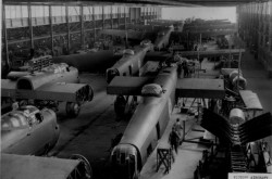 A black and white photograph of approximately 10 airplanes under construction in a large factory. Two workers are visible in the foreground.