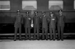 Black and white photograph of 6 uniformed Black porters standing in a line in front of a train. Some porters are smiling and looking at each other.