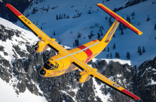 Yellow and red Buffalo CC-115 aircraft in flight, set against a snowy mountainous terrain.