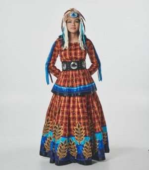 Jeune femme portant un magnifique costume orange et bleu et une coiffe orange et bleue assortie, faite de perles, de cuir et de plumes. Elle a les yeux et les cheveux foncés et se tient debout, les mains derrière le dos.