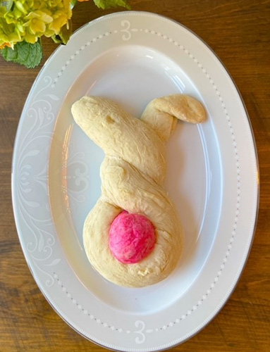 A piece of specialty bread sits on a white, oval plate, on top of a wooden surface. The dough of the bread has been twisted into the approximate shape of a rabbit, and a pink-coloured dough ball represents the tail.