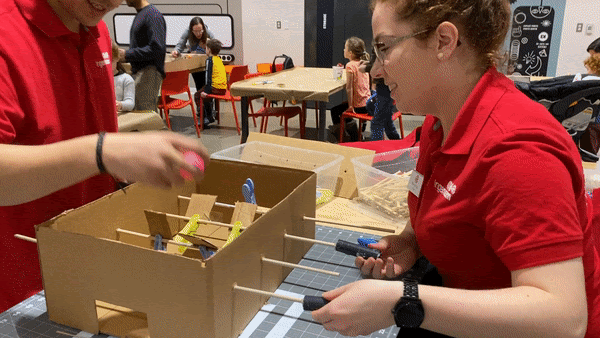 Un jeune homme et une jeune femme en polos rouges d’Ingenium se disputent un match de baby-foot version carton, et l’un d’eux compte un but avec une balle rose. Les joueurs de carton sont retenus à l’aide d’épingles à linge bleues et jaunes. 