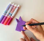 Artist’s hand is colouring the origami rabbit shape on a white surface.   Four coloured markers are also on the white surface.
