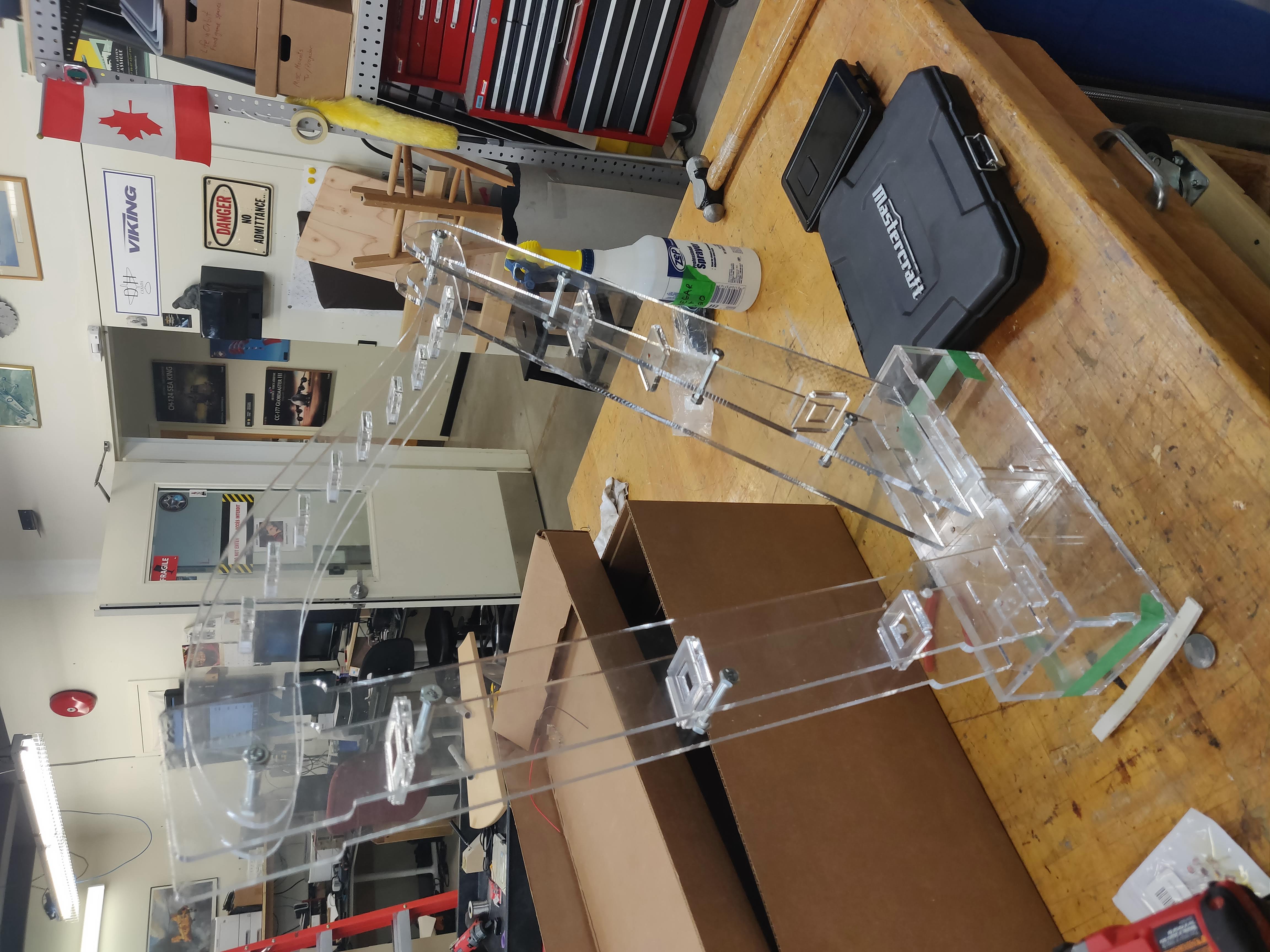 A clear, plastic harp-shaped frame sits on top of a wooden work surface. Tools, boxes, and a computer desk can be seen in the background.