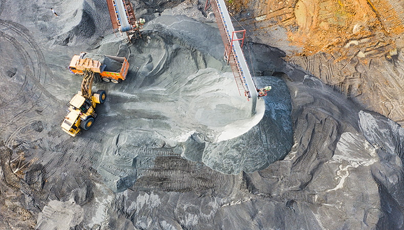From a view far above a yellow excavator dumping grey dust into a yellow dump truck.  Two long thin conveyor belts can also be seen releasing grey dust onto the ground.